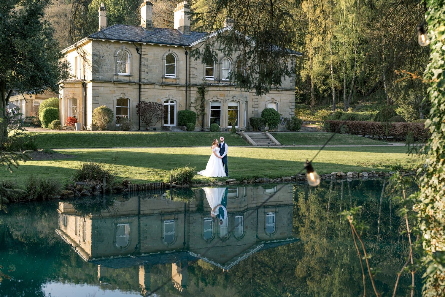 A scenic panoramic view of a historic stone building reflected in a tranquil pond surrounded by autumn trees.