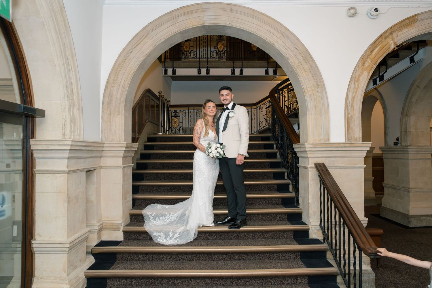Bride and groom posing on elegant stone staircase with arched architecture in a classic wedding venue.