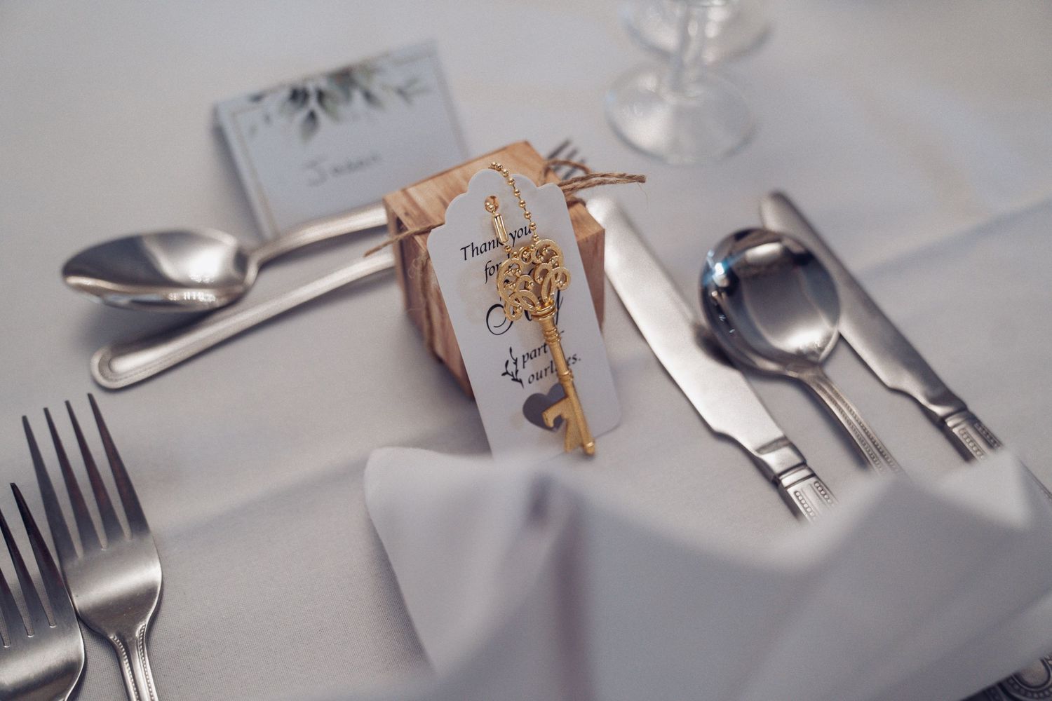 Elegant place setting with silverware and decorative gold-trimmed wedding favor on white tablecloth.