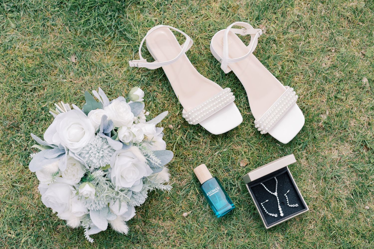 White bridal accessories including sandals, floral bouquet, small blue bottle and black ring box arranged on grass.
