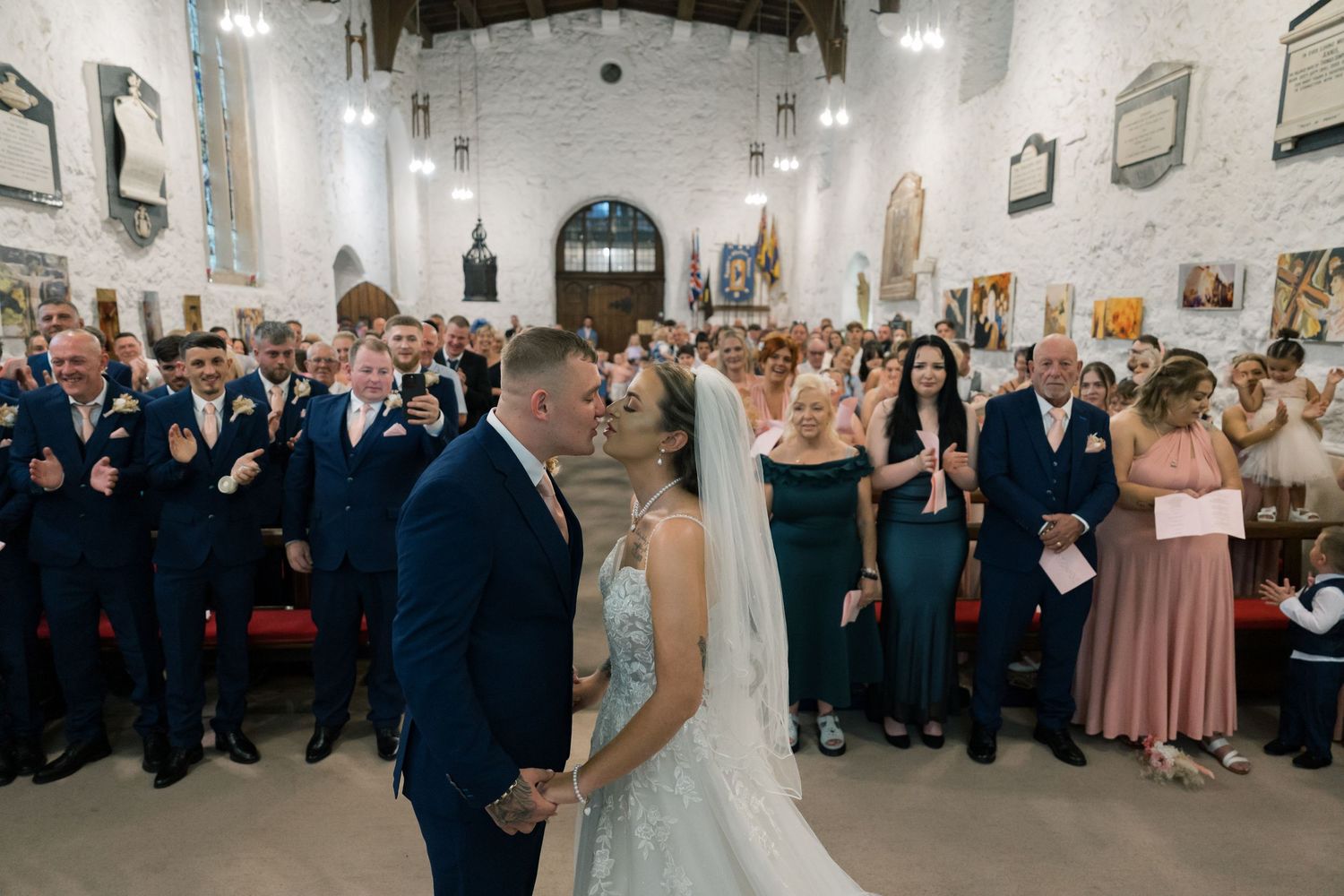 Wedding guests celebrate with raised arms on the dance floor of a rustic chapel decorated with twinkling lights.