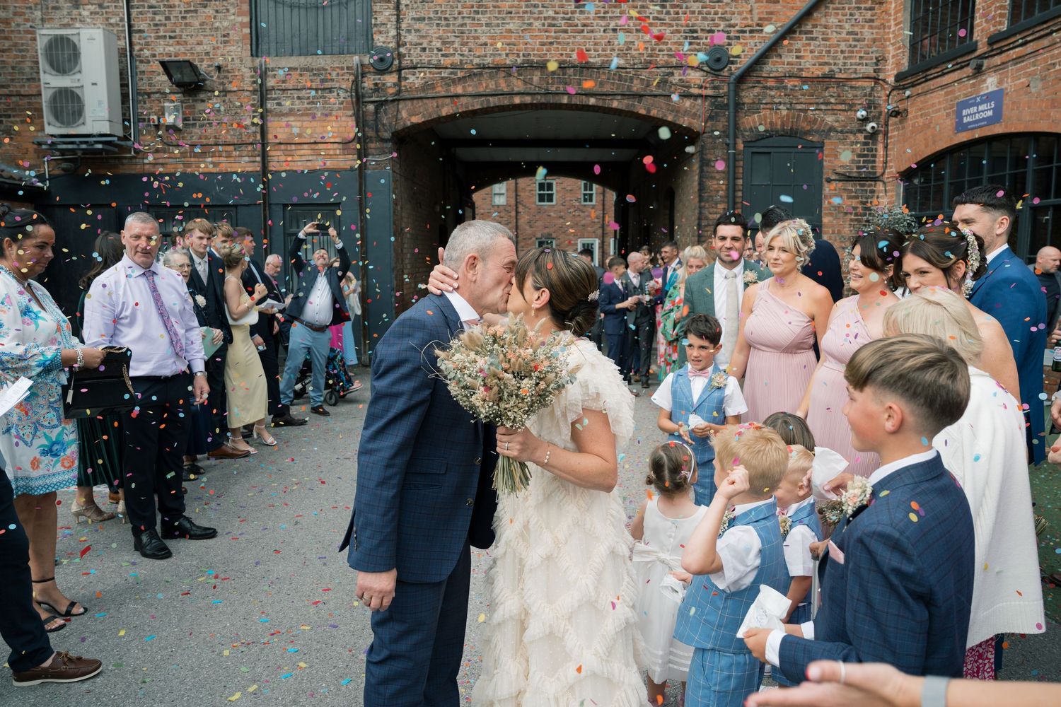 Wedding celebration outside a brick building with guests throwing confetti.