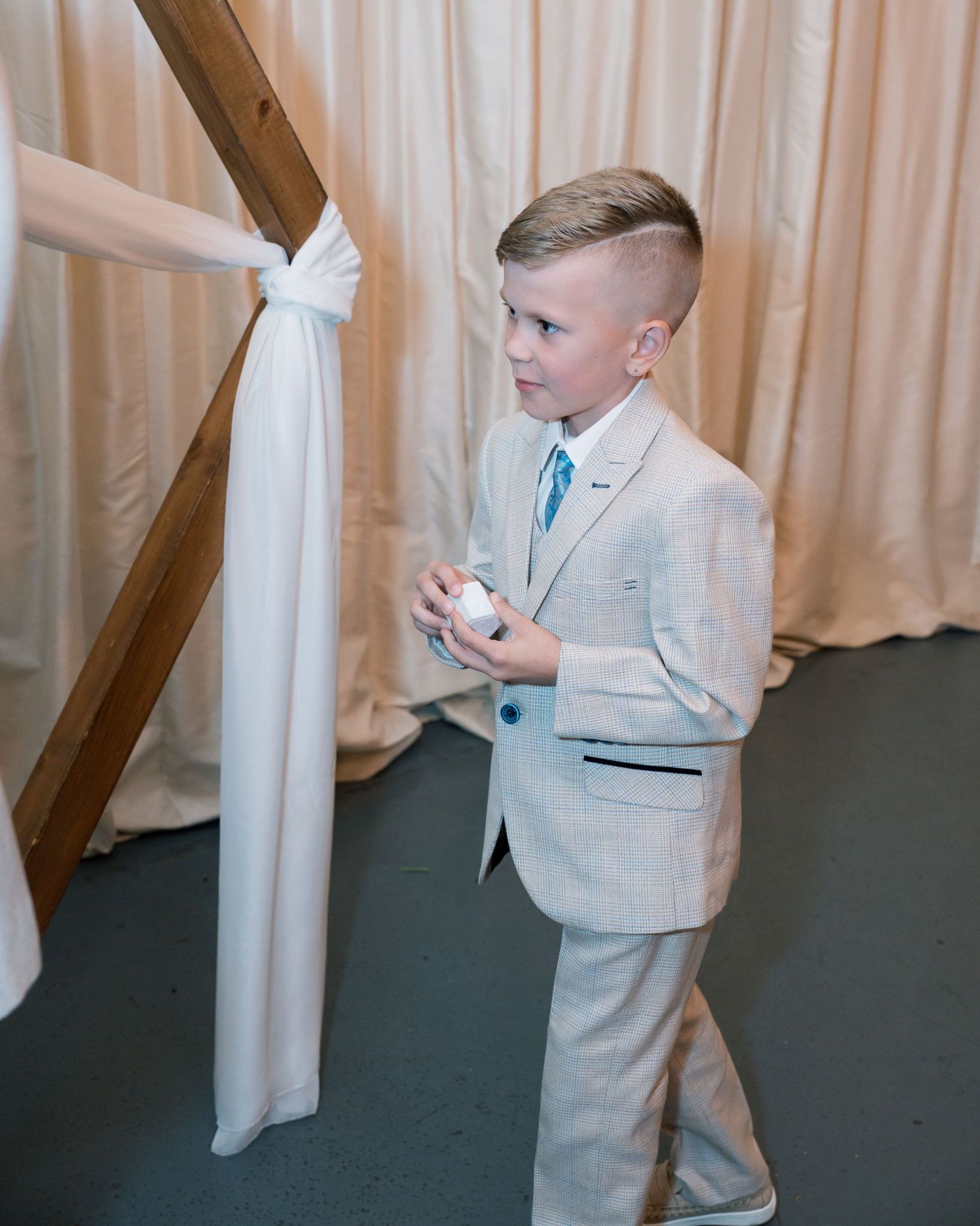 Young ring bearer in a light-colored suit standing by white curtains at a wedding ceremony.