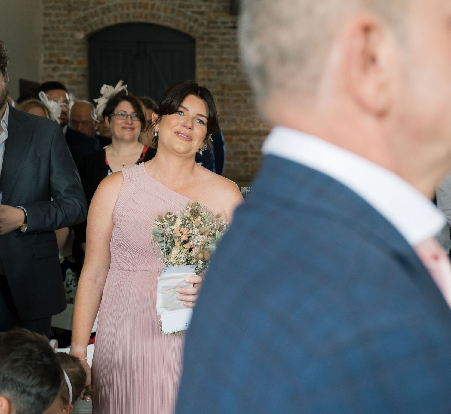 A group celebrates at a wedding reception in a rustic brick-walled venue with soft lighting and pink bridesmaid dresses.