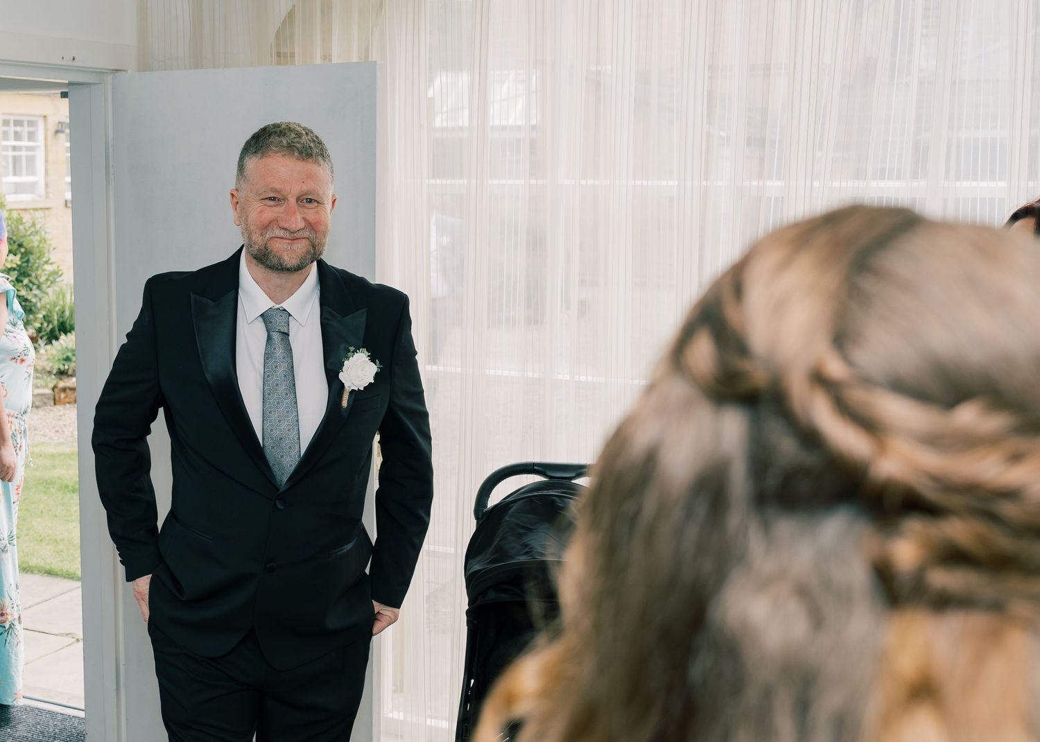 A person in a black suit stands in an elegant room with large windows and natural lighting.