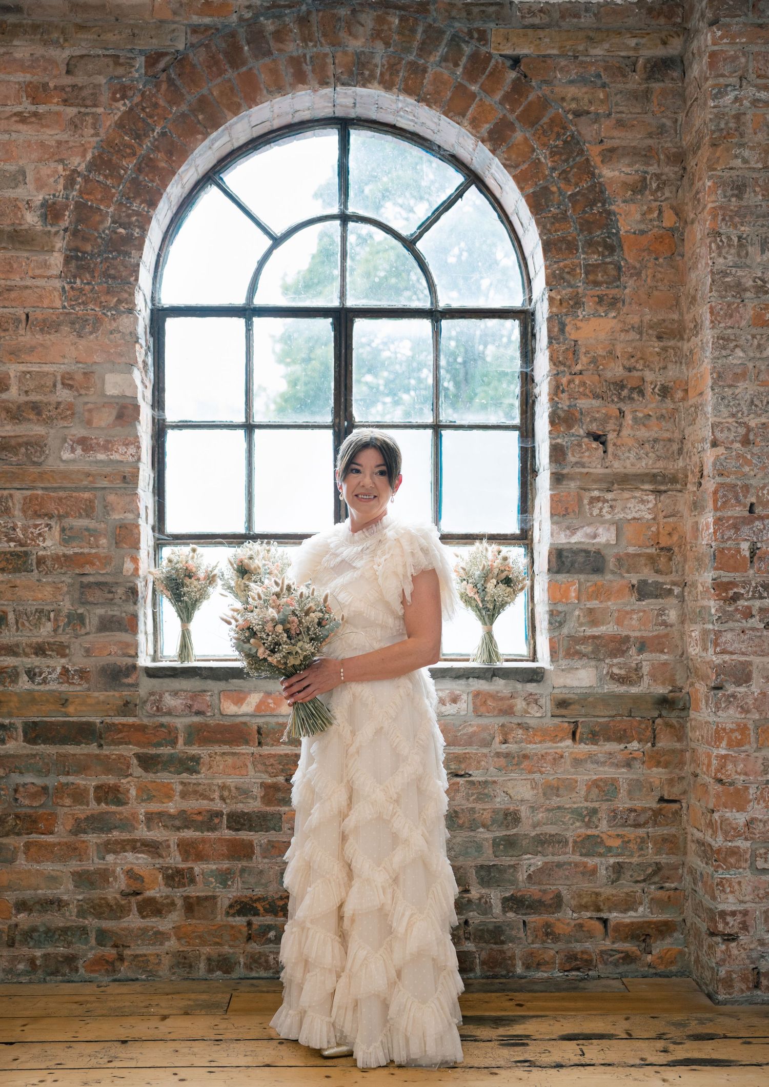 Elegant ruffled white wedding dress photographed against rustic brick walls with arched windows in an industrial venue.