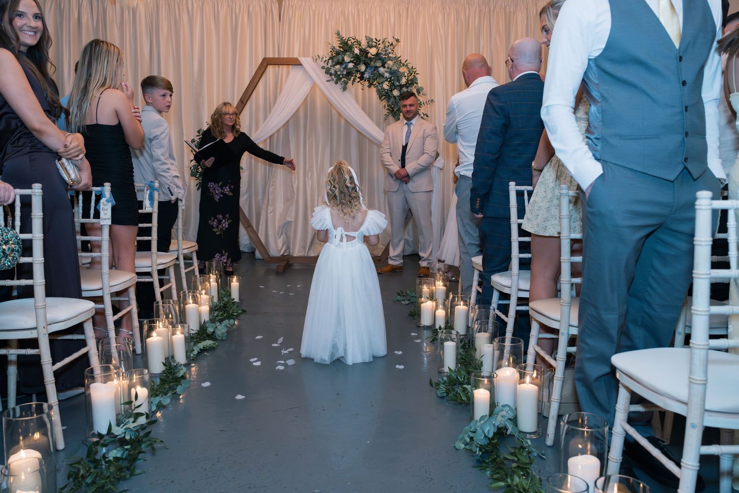 A flower girl walks down an aisle lined with glowing lanterns and white chairs at an indoor wedding ceremony.