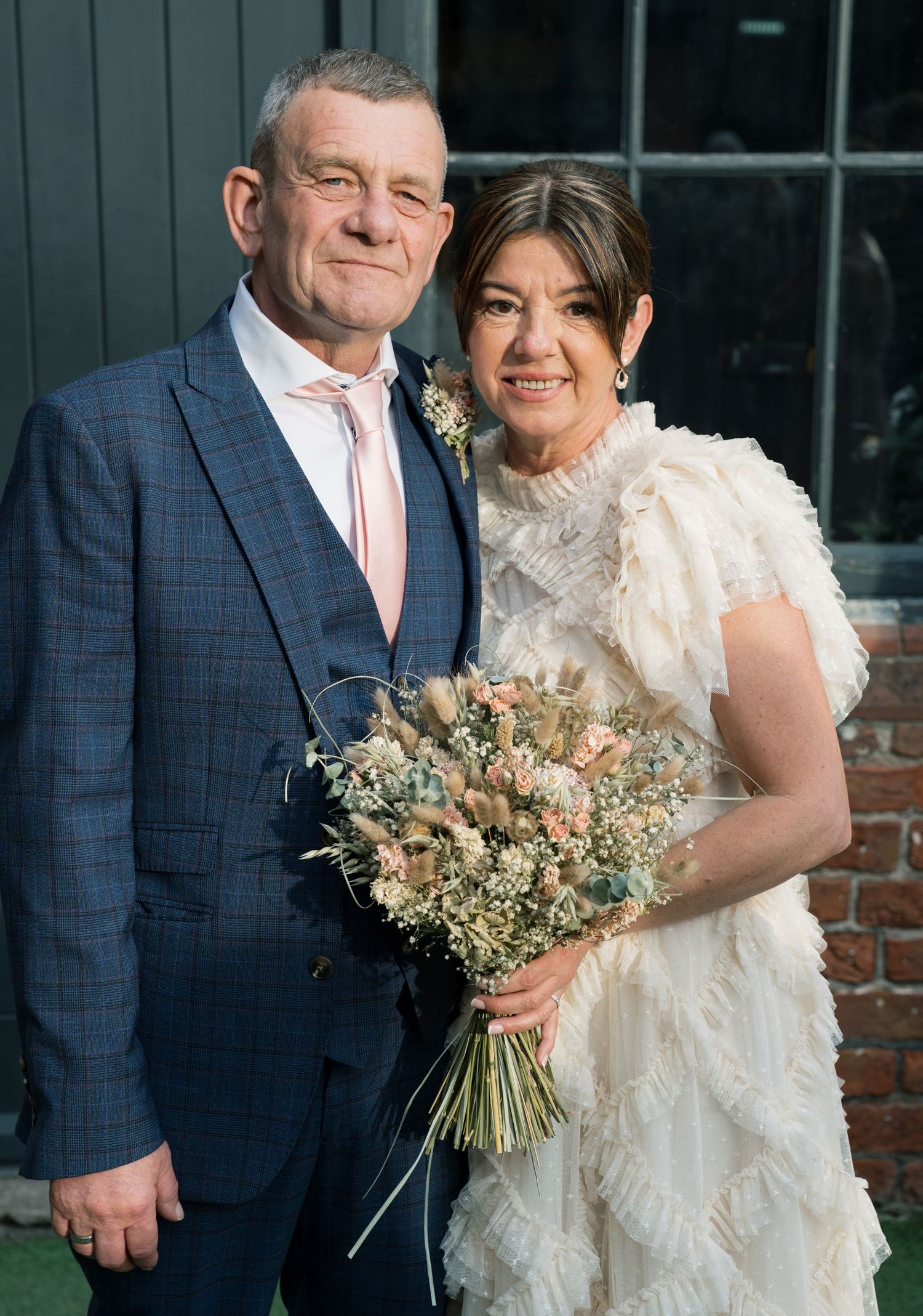 A couple in wedding attire pose together, with the bride holding a dried flower bouquet.