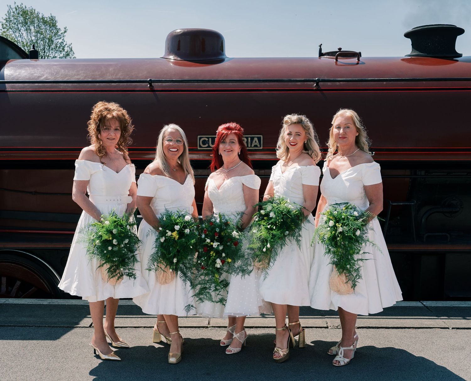 Five bridesmaids in white dresses sit on a bench holding green bouquets during a wedding photo session.