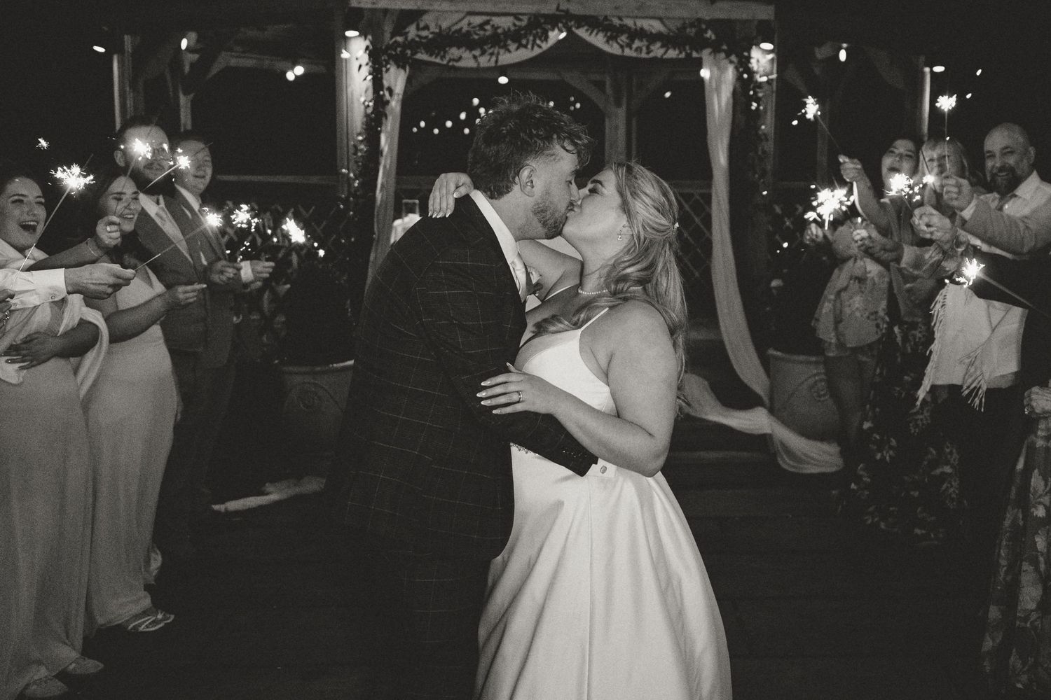 Black and white photo of bride and groom kissing surrounded by guests holding sparklers at night, at Whitley Hall, Sheffield.