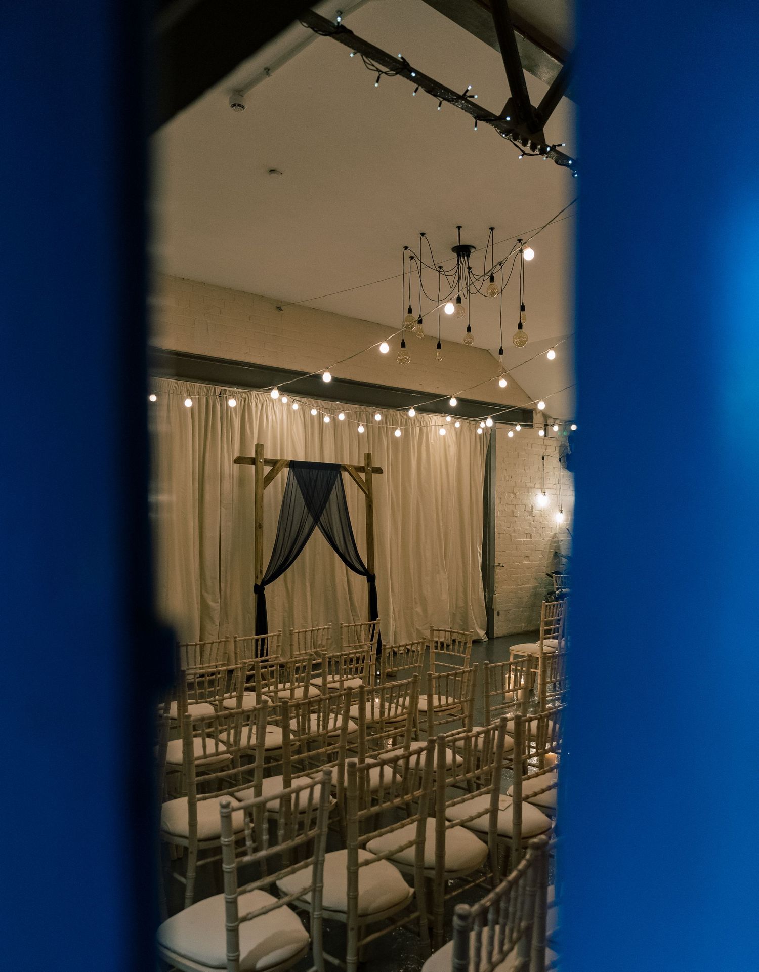 Wedding ceremony setup with draped arch and chiavari chairs under string lights.