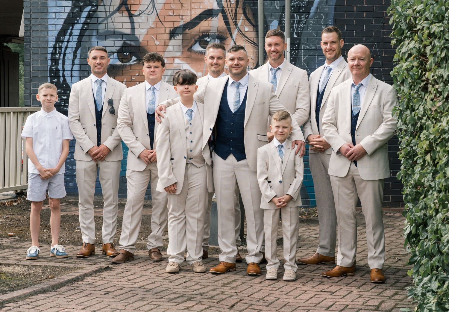 A wedding party in light beige and white attire poses together on a brick pathway outdoors.