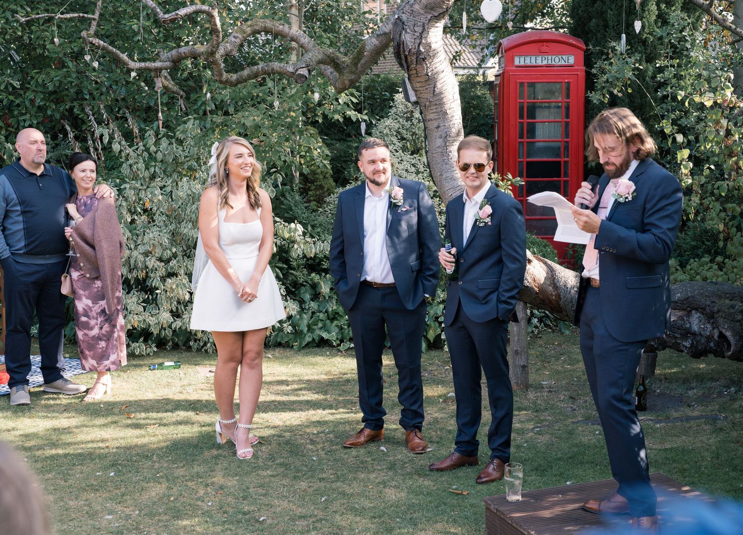 Outdoor wedding celebration with red phone booth visible among trees and guests.