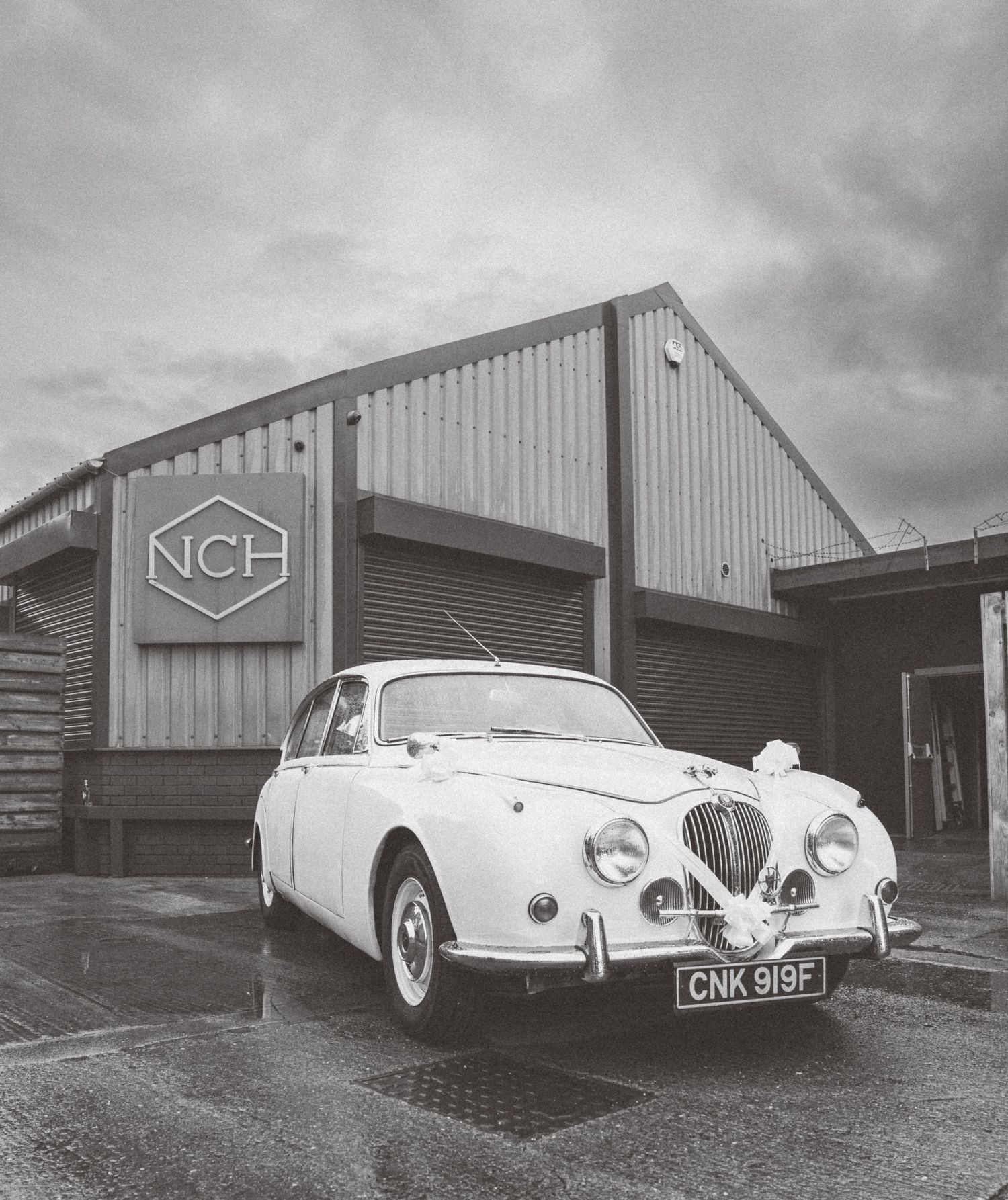 Classic white vintage car parked outside industrial metal buildings with NCH signage.