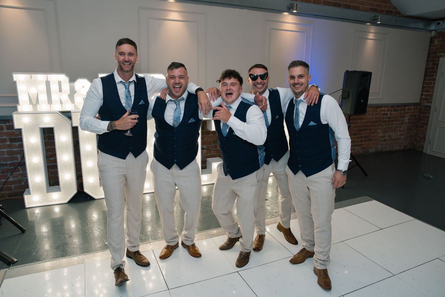 Wedding party in matching navy vests and khaki pants posing by illuminated MR & MRS sign.