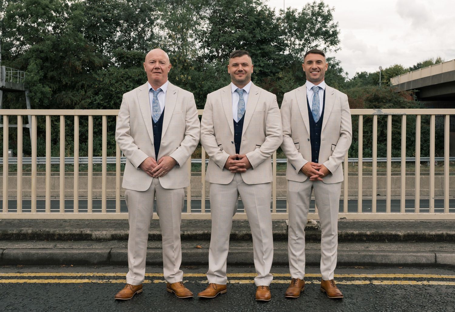 Three groomsmen in matching light beige suits and blue ties pose together on a bridge railing.