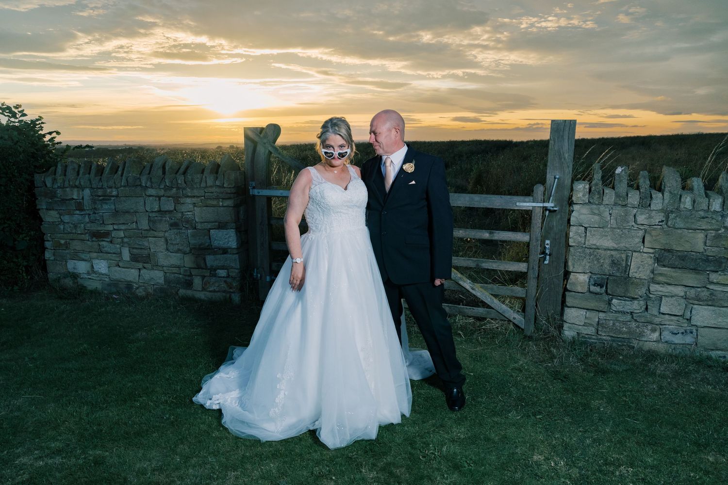 Sunset wedding portrait against a rustic stone wall and wooden gate in the countryside.