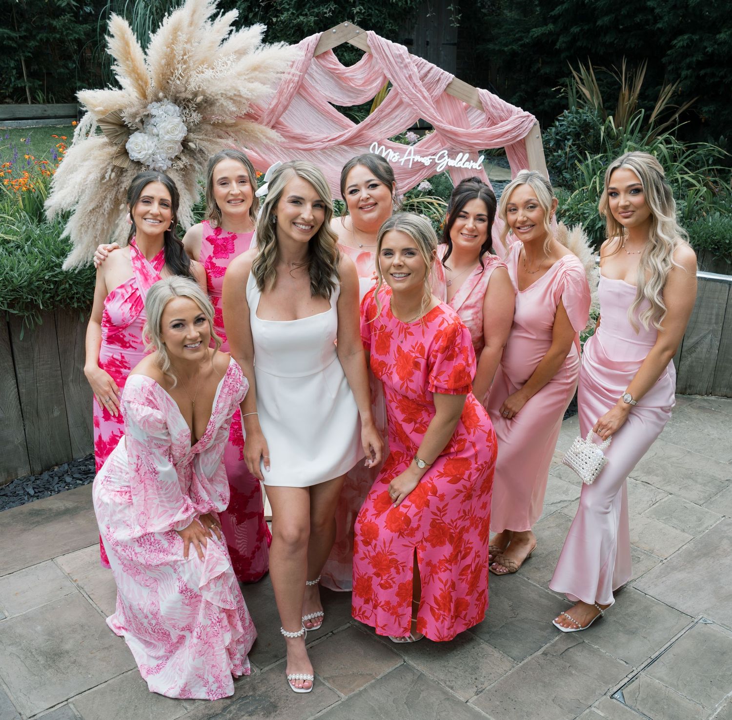 A group in coordinating pink and coral floral dresses poses together outdoors during a special occasion.