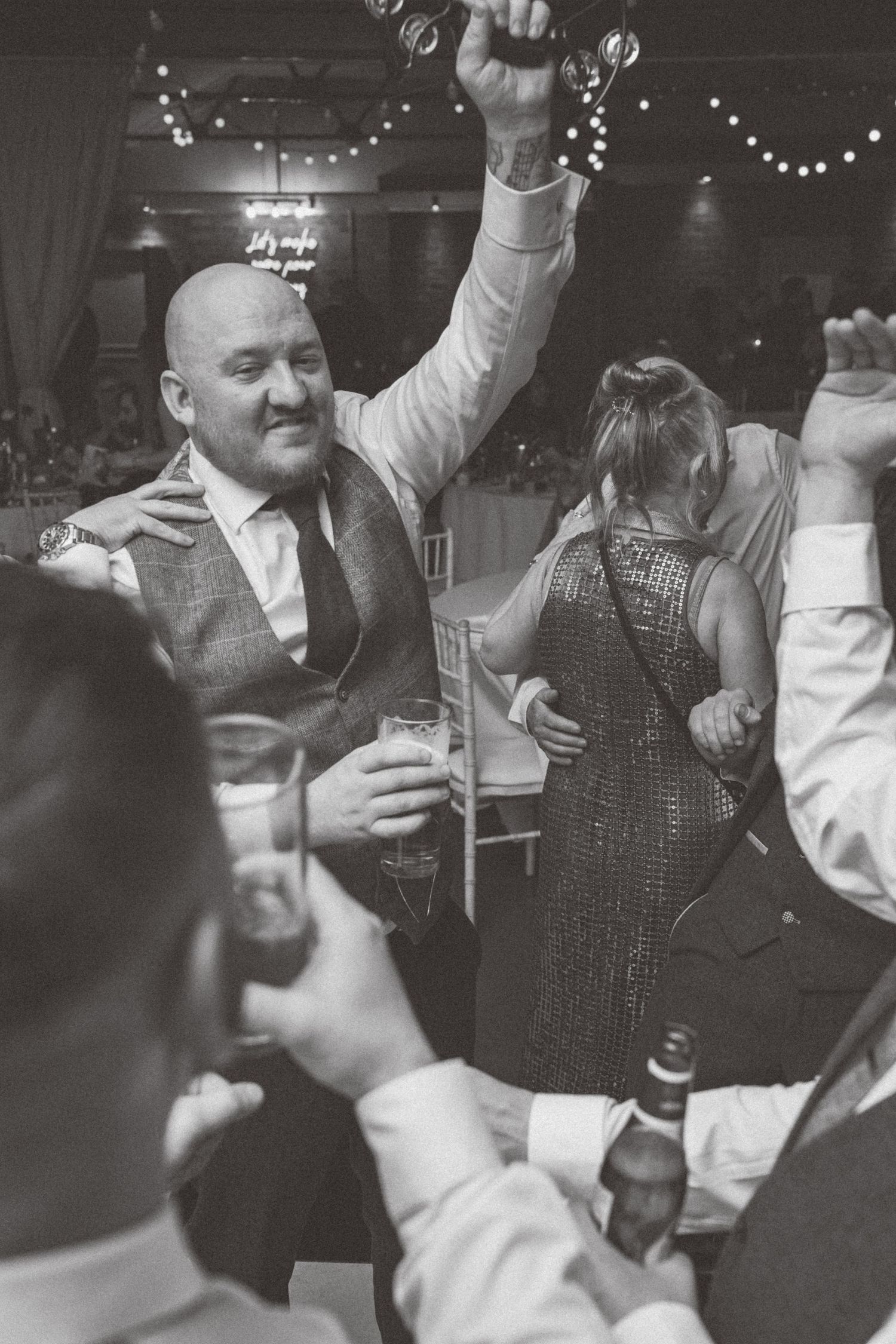 Black and white photo of wedding guests celebrating and dancing with arms raised at a reception.