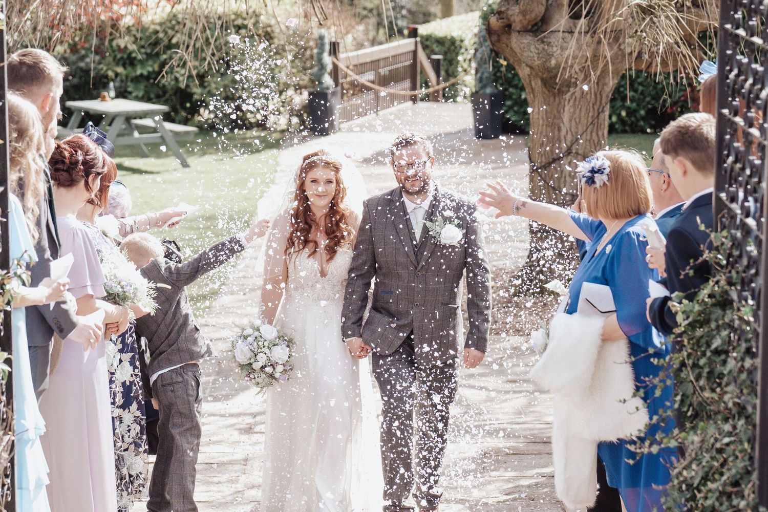 Wedding guests showering the newlyweds with confetti along a garden path.