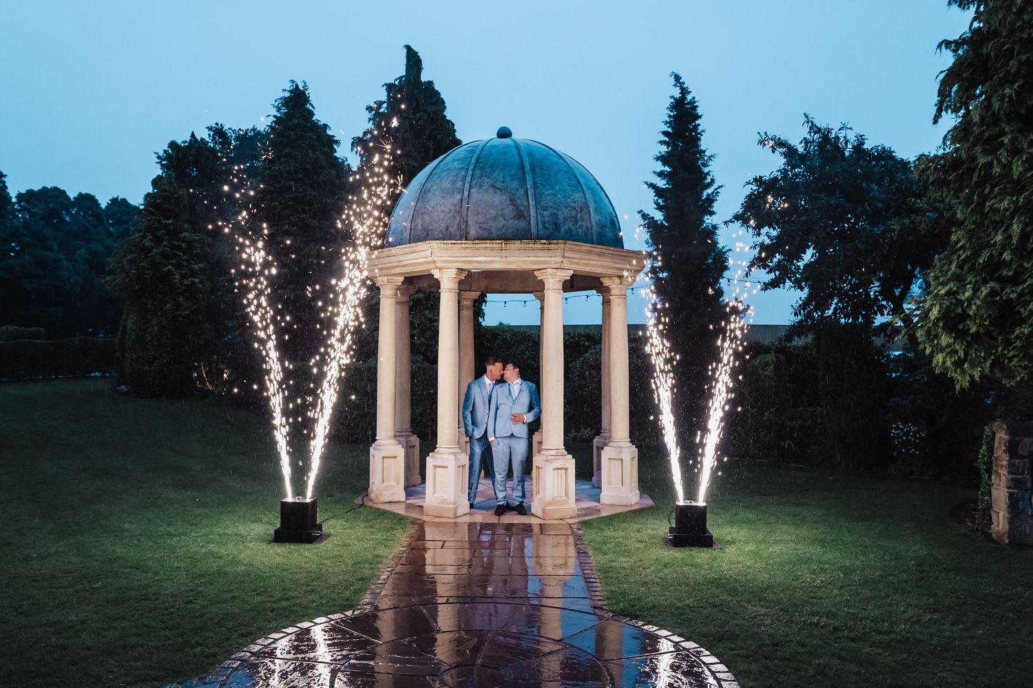 A domed gazebo lit up with sparklers at dusk creates a romantic outdoor atmosphere in a garden setting.
