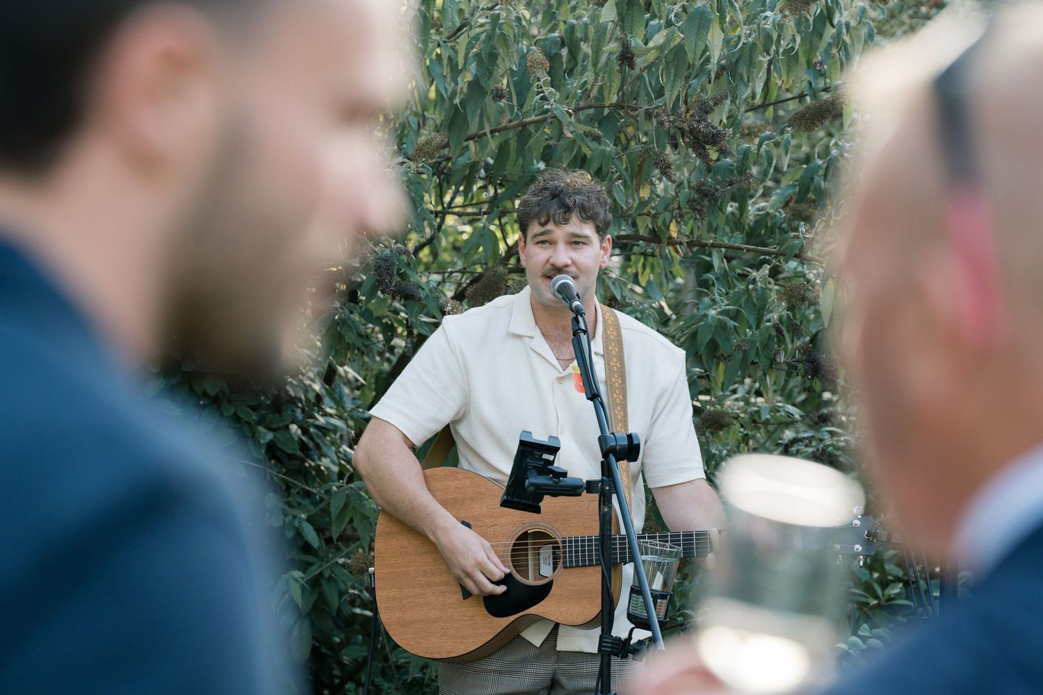 A musician plays acoustic guitar at an outdoor wedding reception surrounded by greenery.