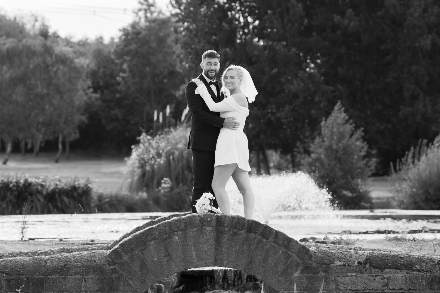 Black and white wedding photo of bride and groom embracing on stone bridge surrounded by lush trees.