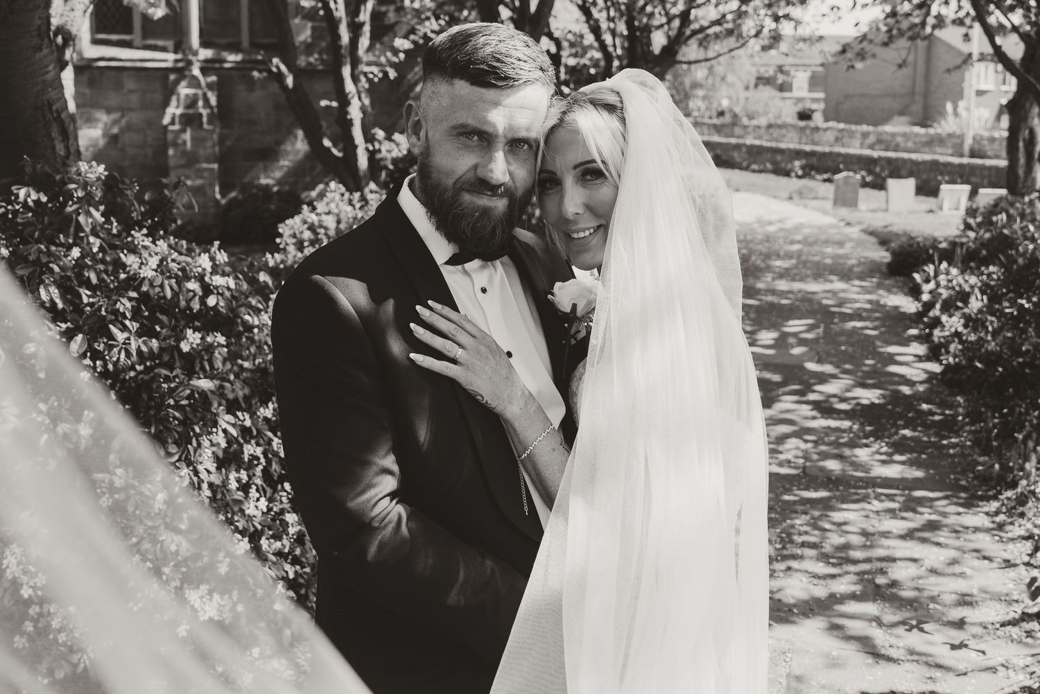 Black and white wedding photo of bearded groom in tuxedo and bride in veil embracing outdoors.