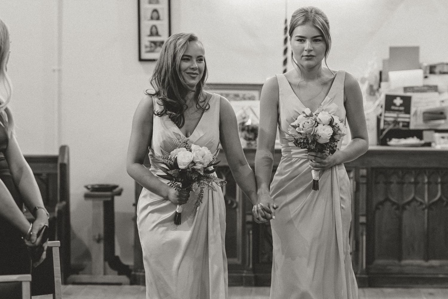 Two bridesmaids in elegant gowns walk down the aisle holding floral bouquets during a wedding ceremony taking place in Kippax, Leeds.