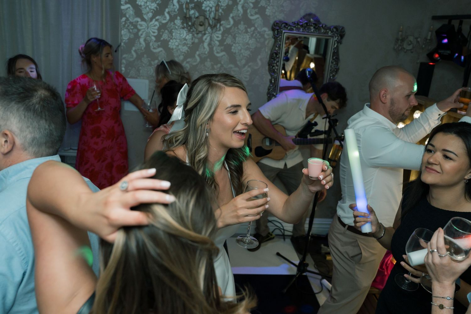 Wedding guests dance and celebrate on crowded dance floor with colorful lighting effects.