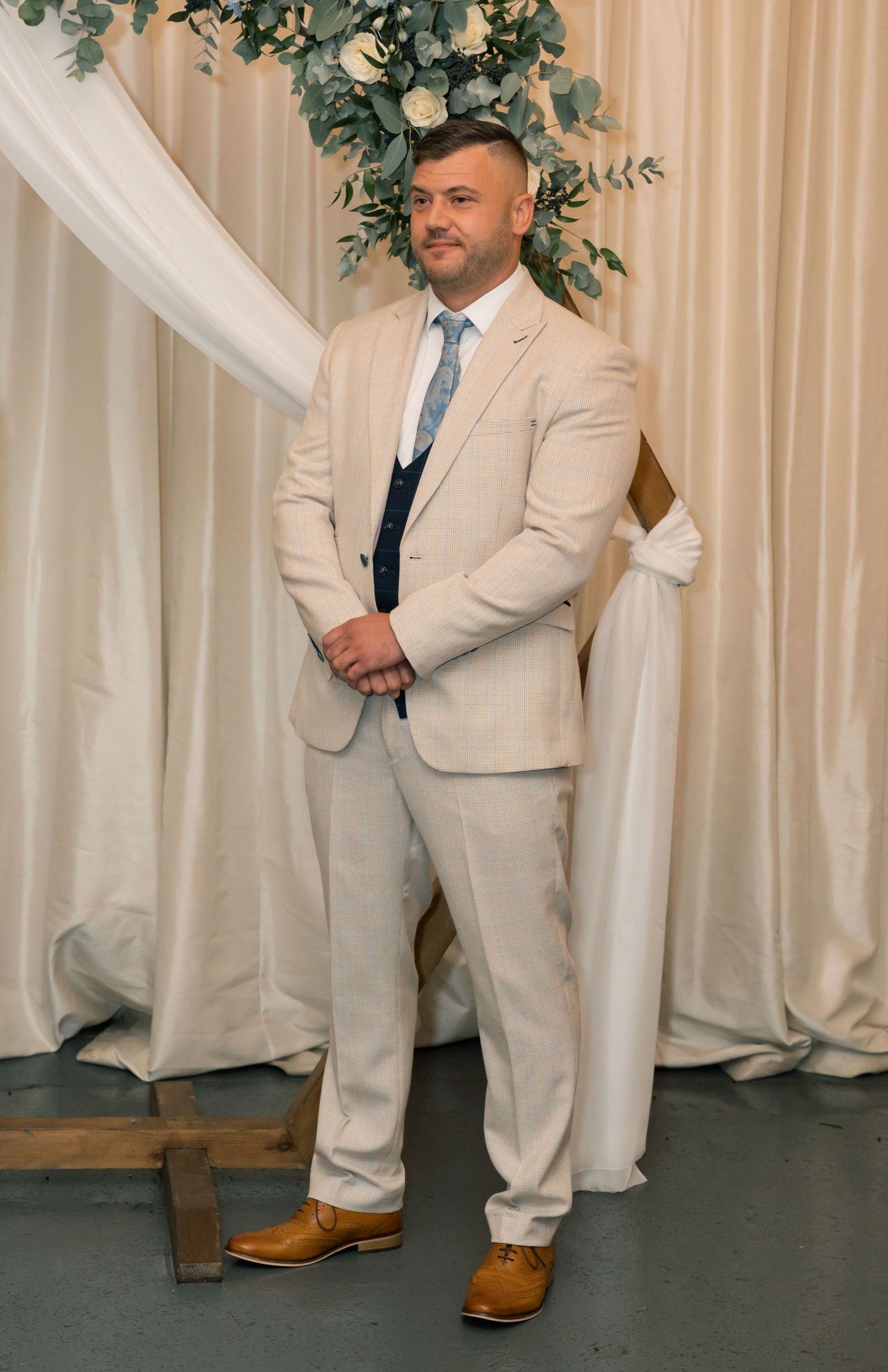 Groomsmen in light beige suits and brown shoes standing against white drapes with floral arrangements.