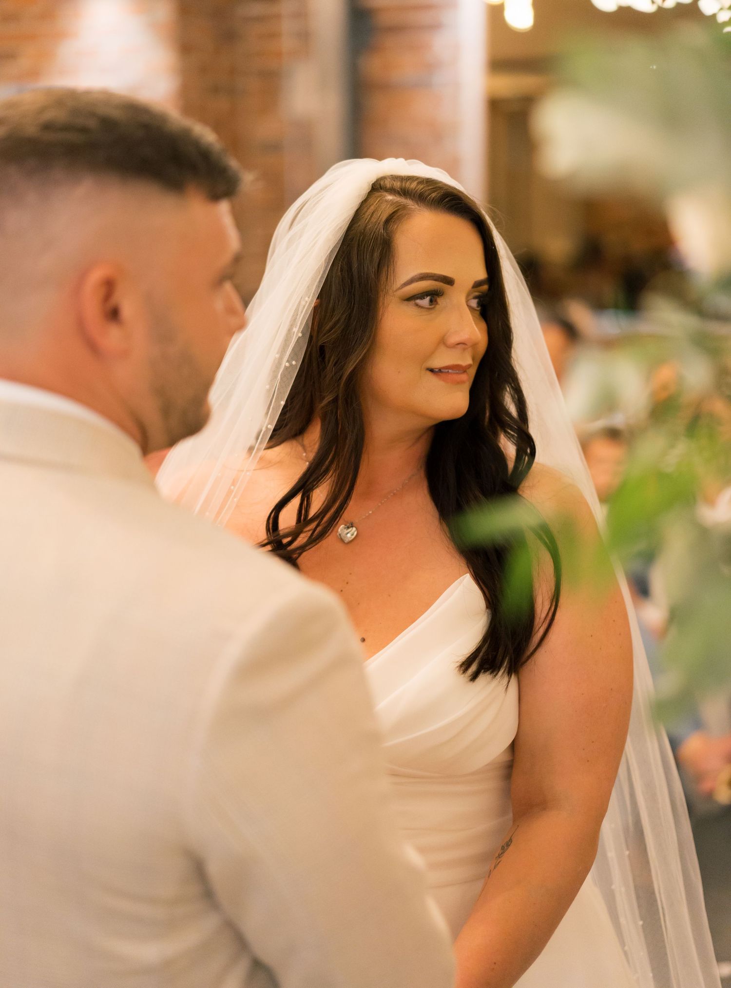 Bride in white dress smiles during wedding reception in brick-walled venue.