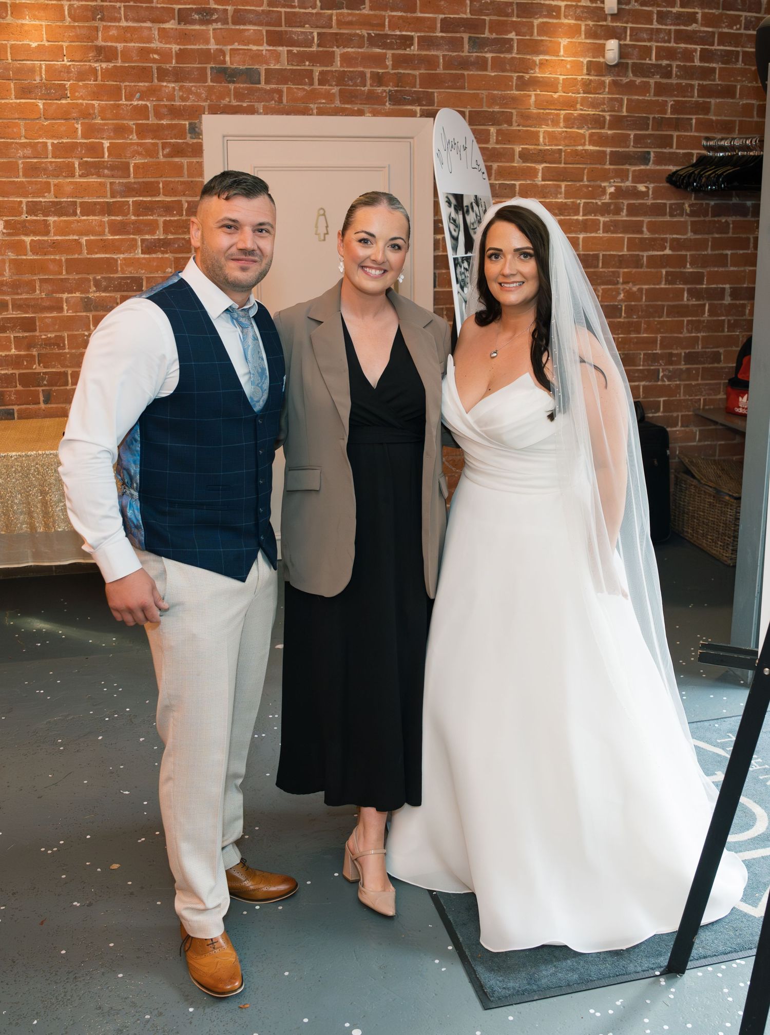Wedding party members pose against a rustic brick wall in formal attire at an indoor venue.