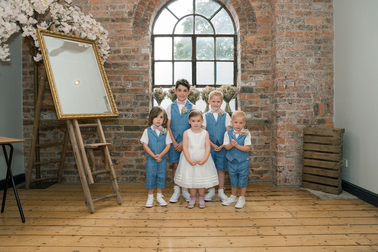 Children in blue outfits sit on wooden floor against exposed brick wall with arched window.