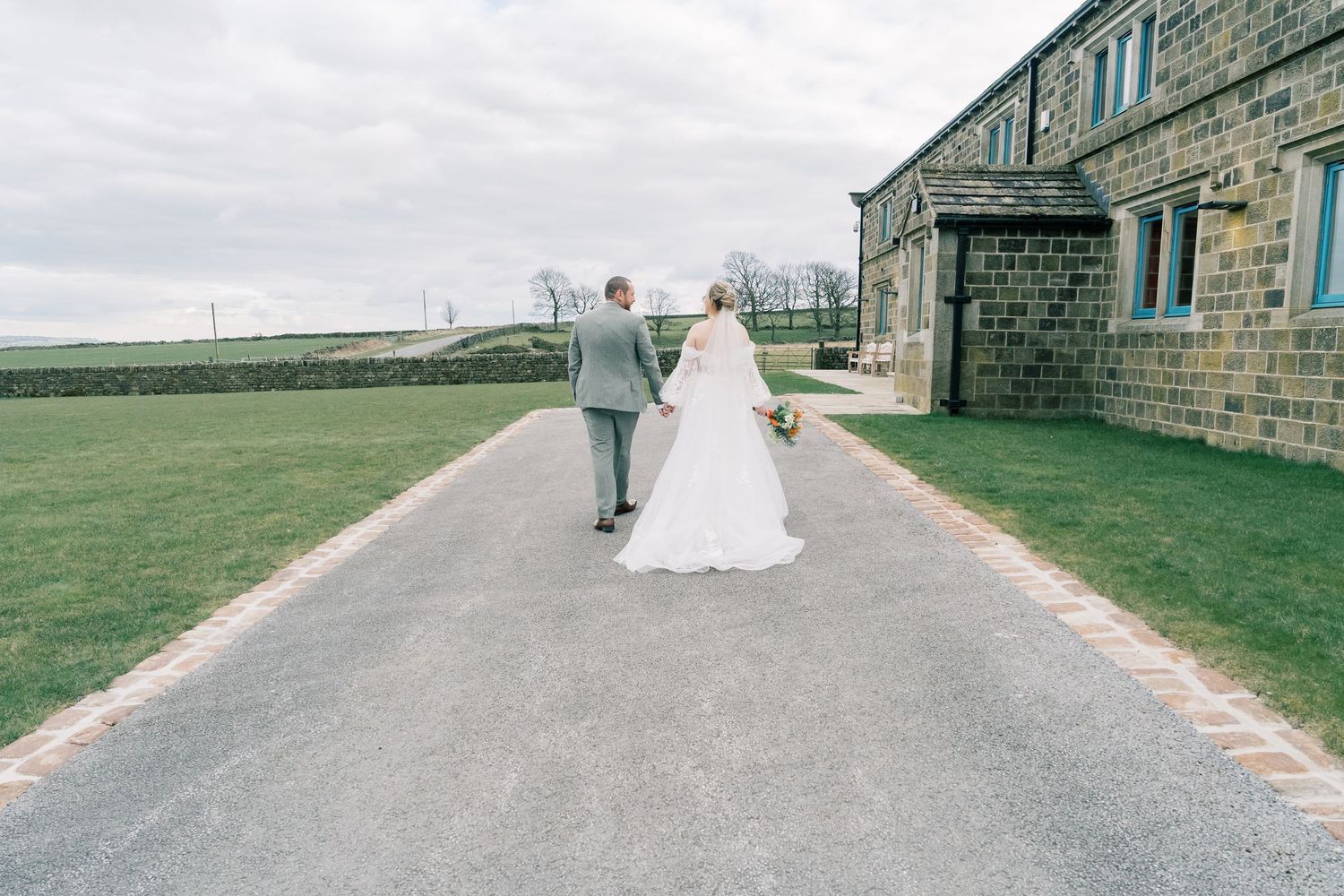 Newlyweds walking down a path beside a rustic stone building on their wedding day.