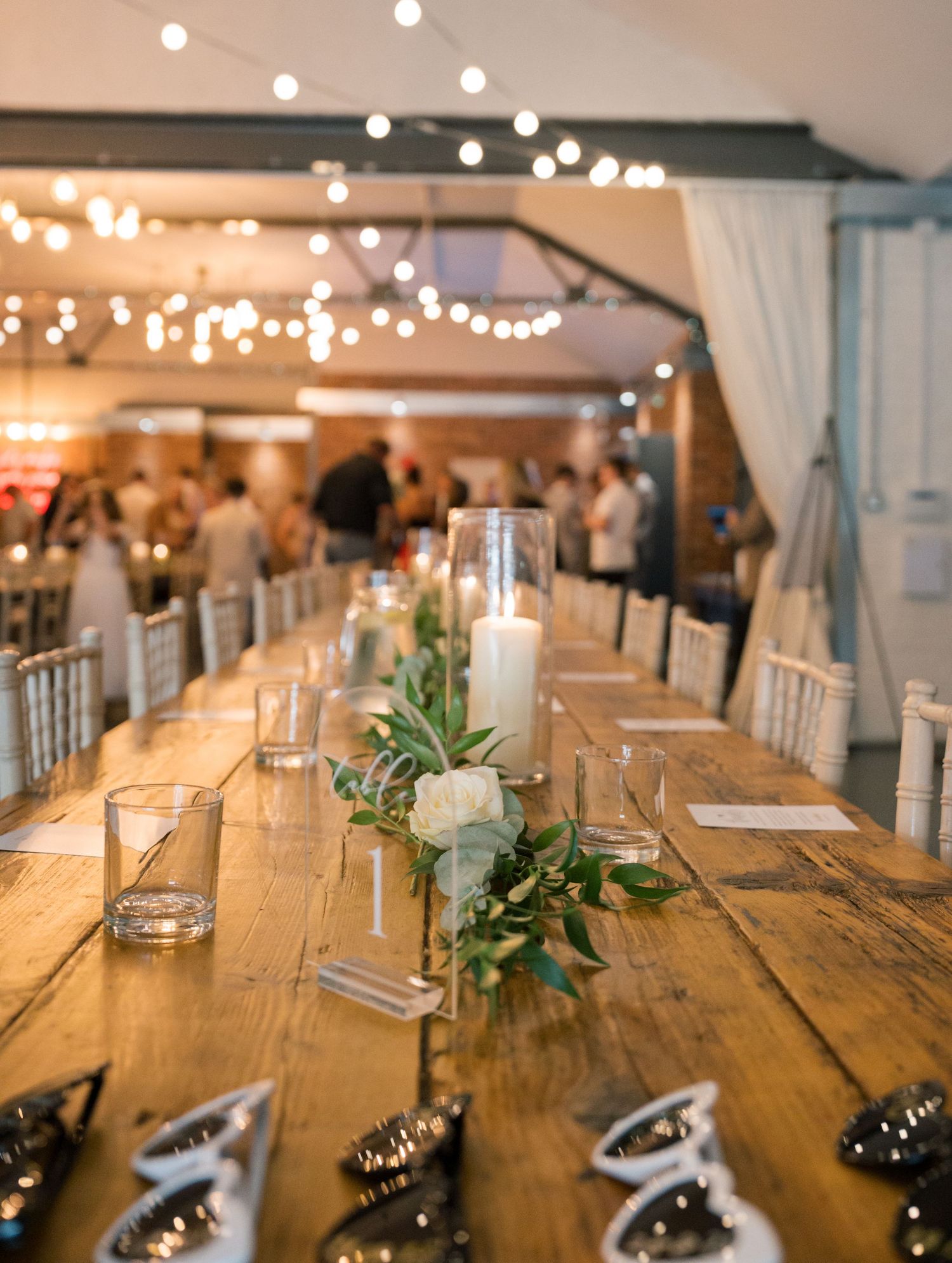 Long wooden tables set for a wedding reception with string lights and greenery decorations.