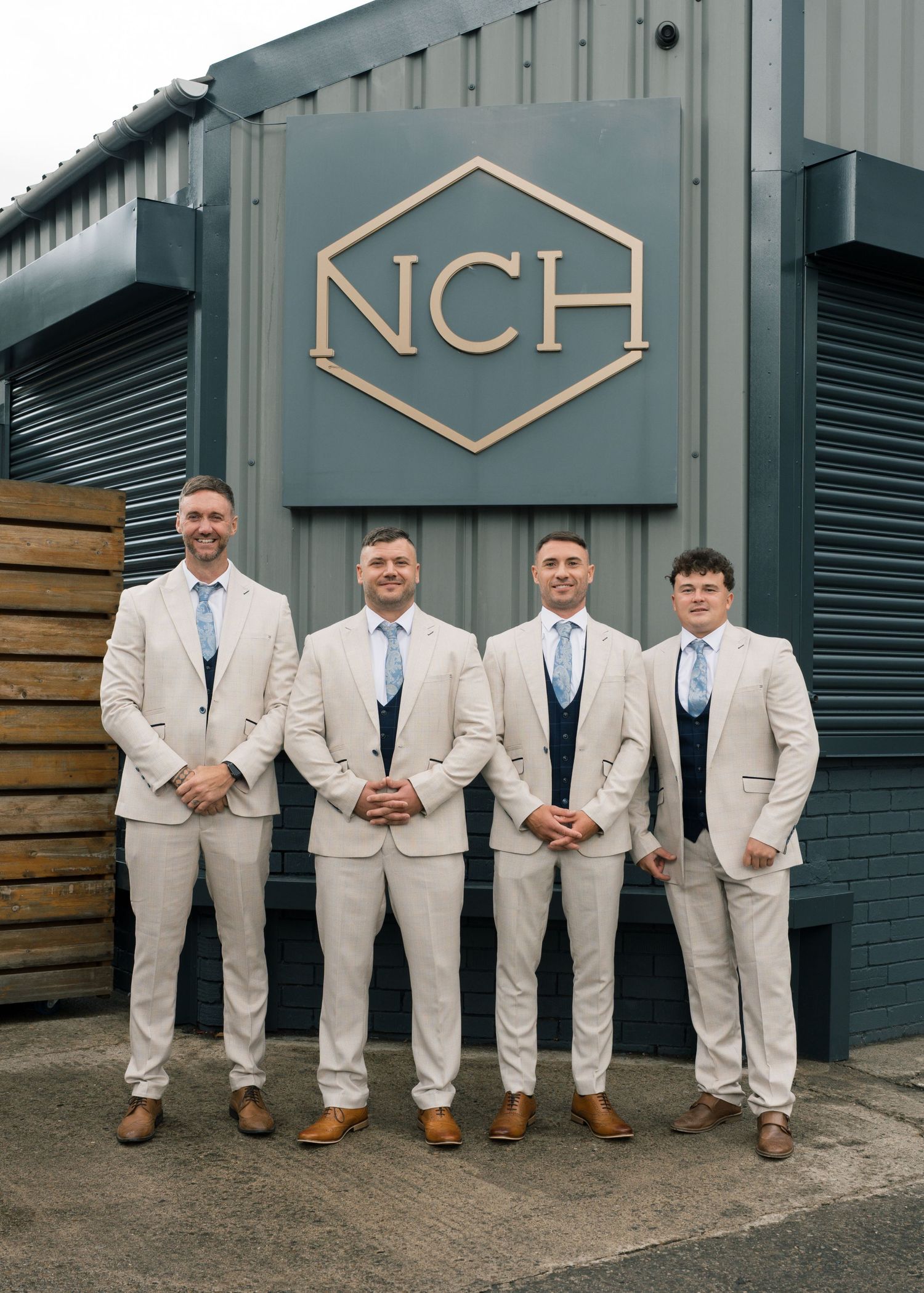 Groomsmen in light beige suits and brown shoes pose casually against an industrial building wall.