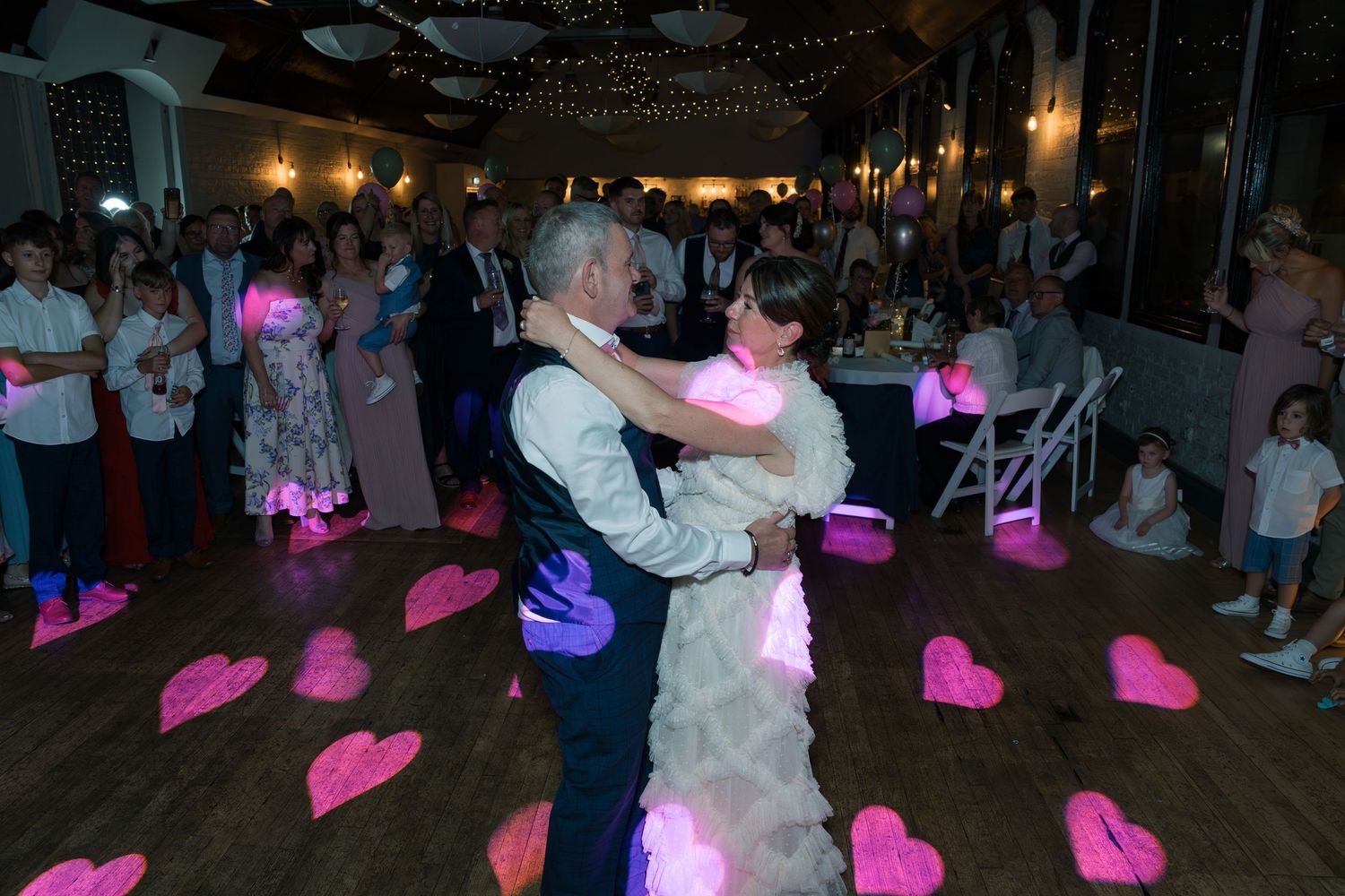 Couple dancing at wedding reception with pink heart projections on dark dance floor.