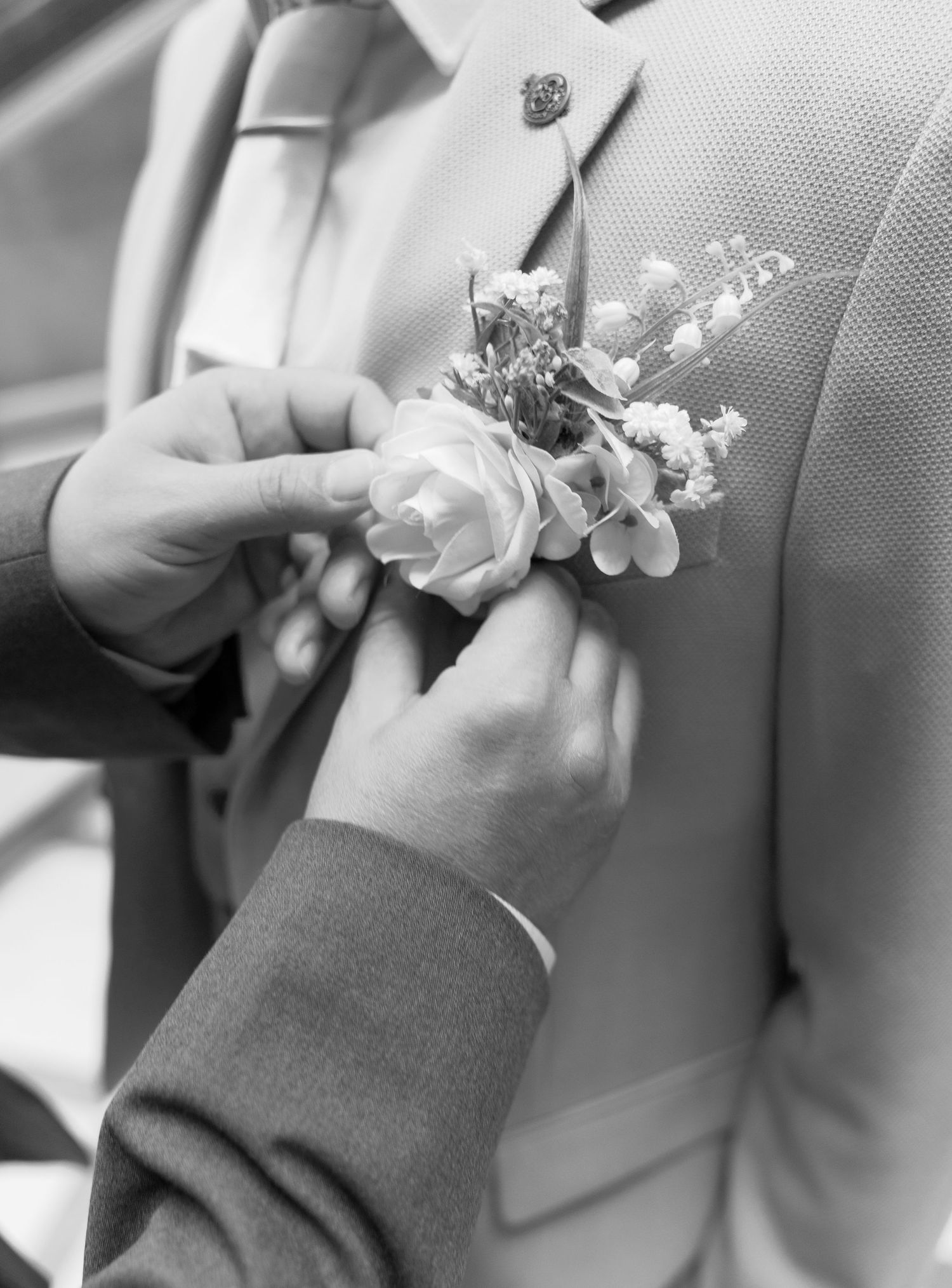 Black and white photo of hands pinning white rose boutonniere on formal suit lapel.