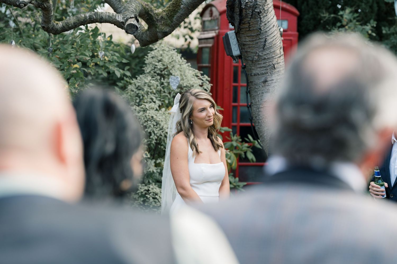 A wedding ceremony scene with a red phone booth visible in the background among trees.