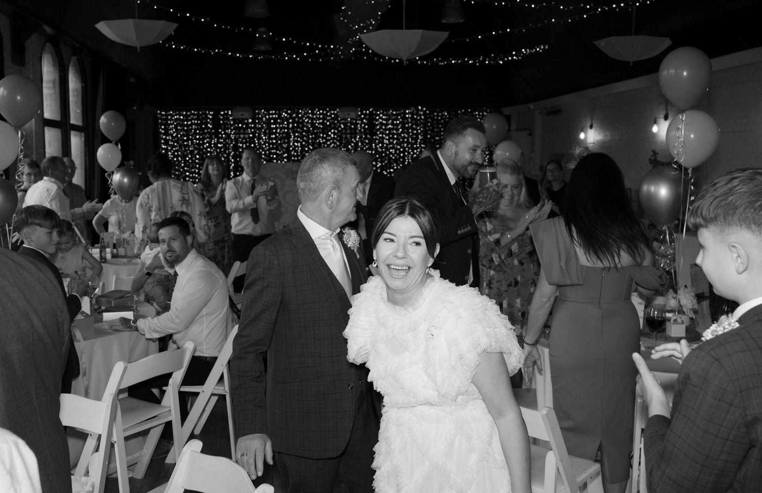Black and white photo of wedding reception with guests seated at tables under string lights.