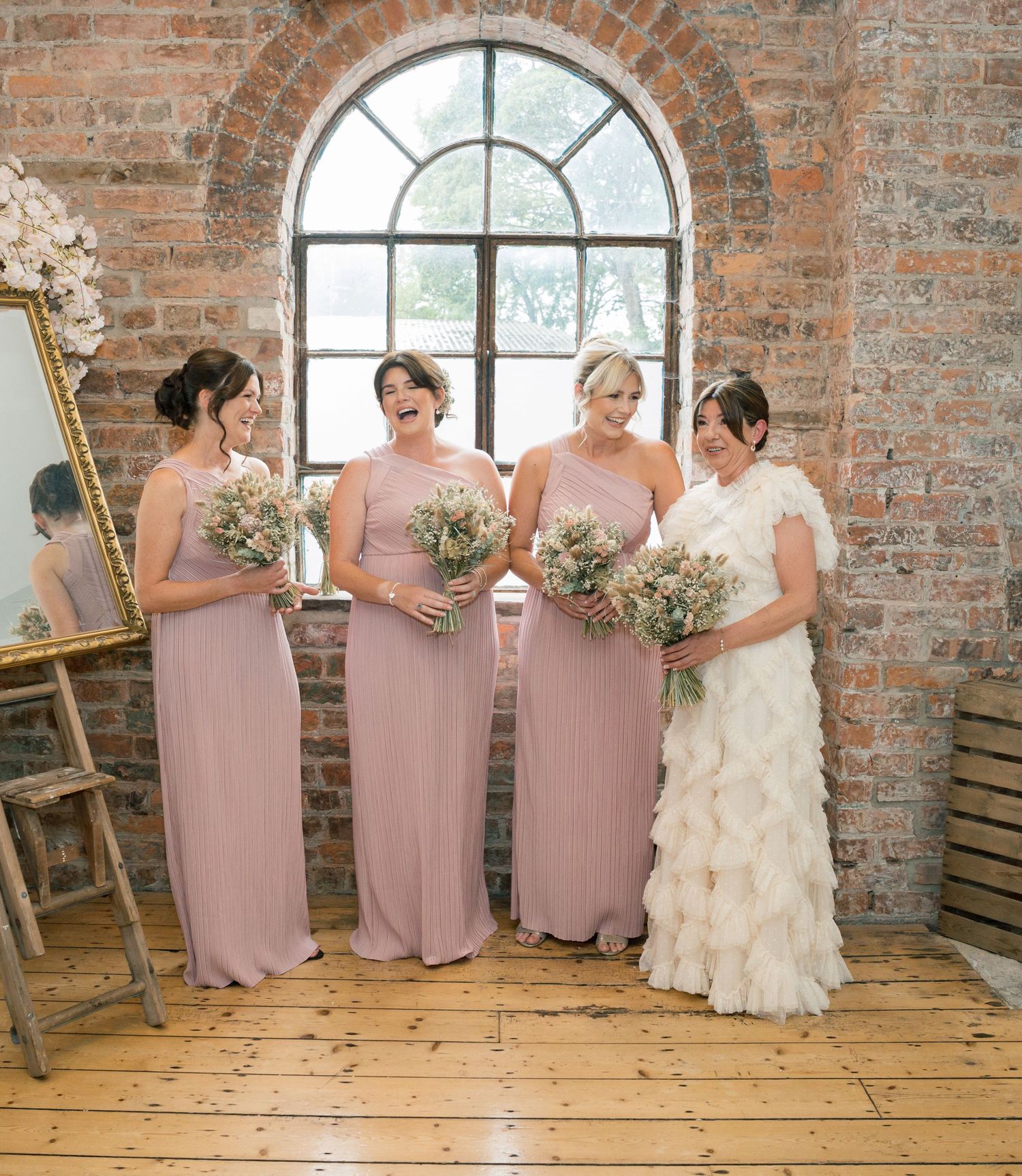Wedding party in blush pink dresses posing by arched window in rustic brick venue.
