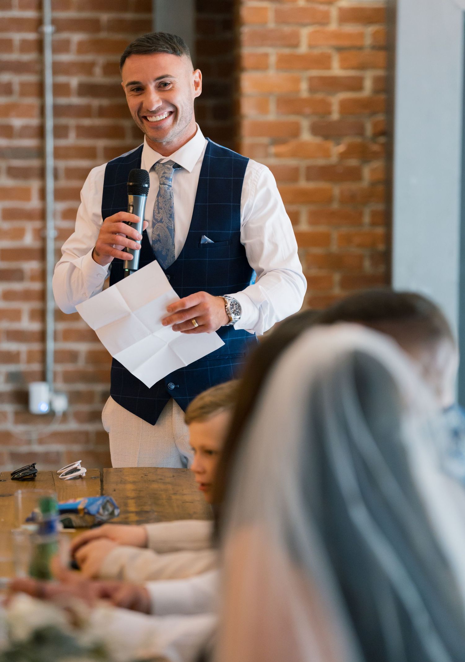 Wedding reception speech being given at rustic brick venue with table settings visible.