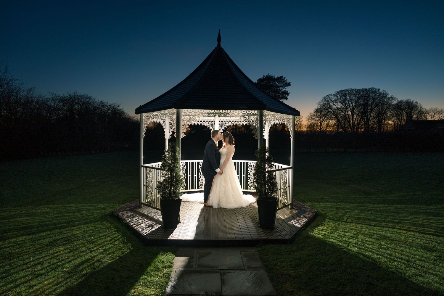 A romantic evening wedding photo of a couple silhouetted in an illuminated white gazebo against a dusky sky, The Bridge Hotel & SPA, Selby
