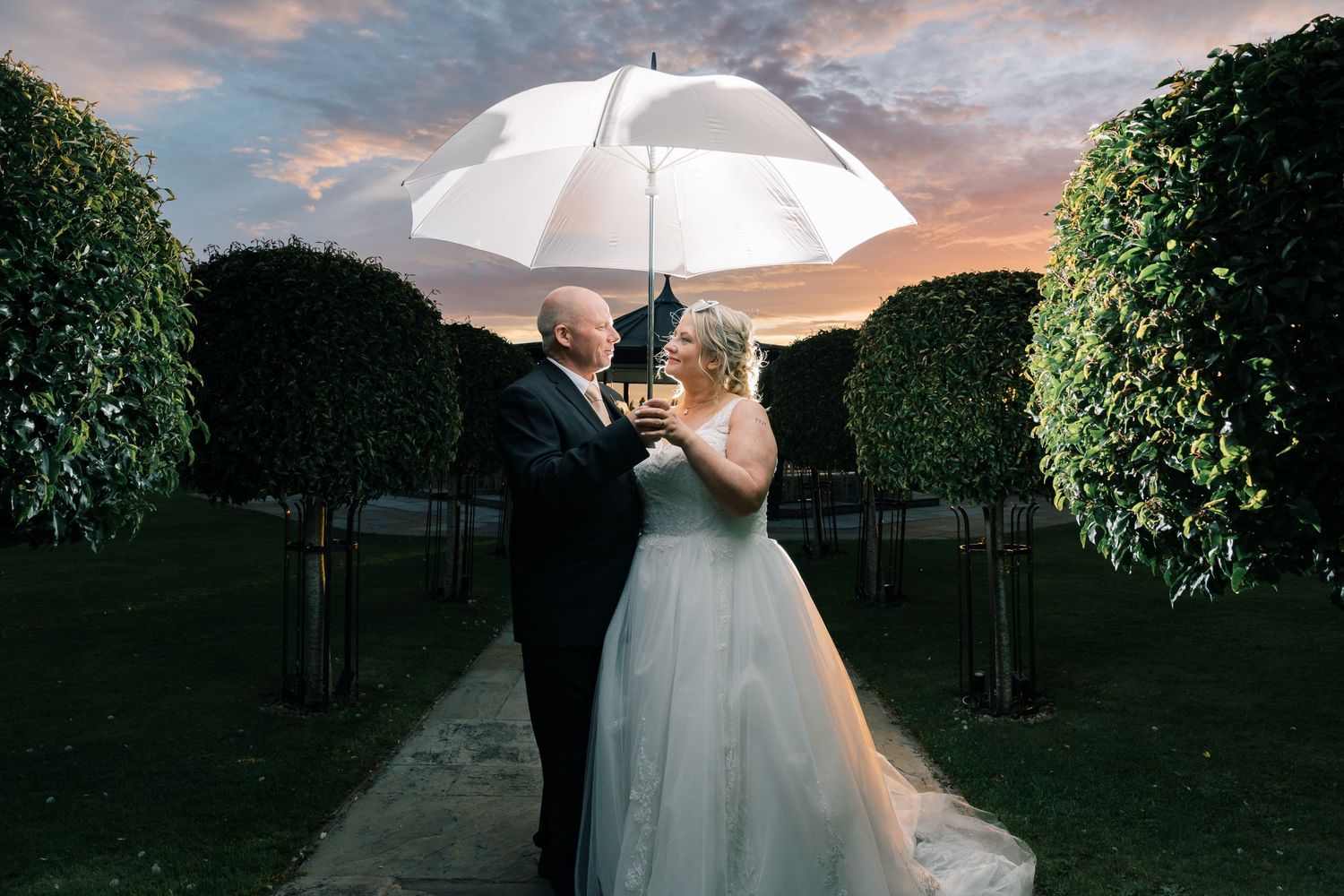 Dramatic sunset wedding photo with couple sharing a white umbrella in a formal garden setting.