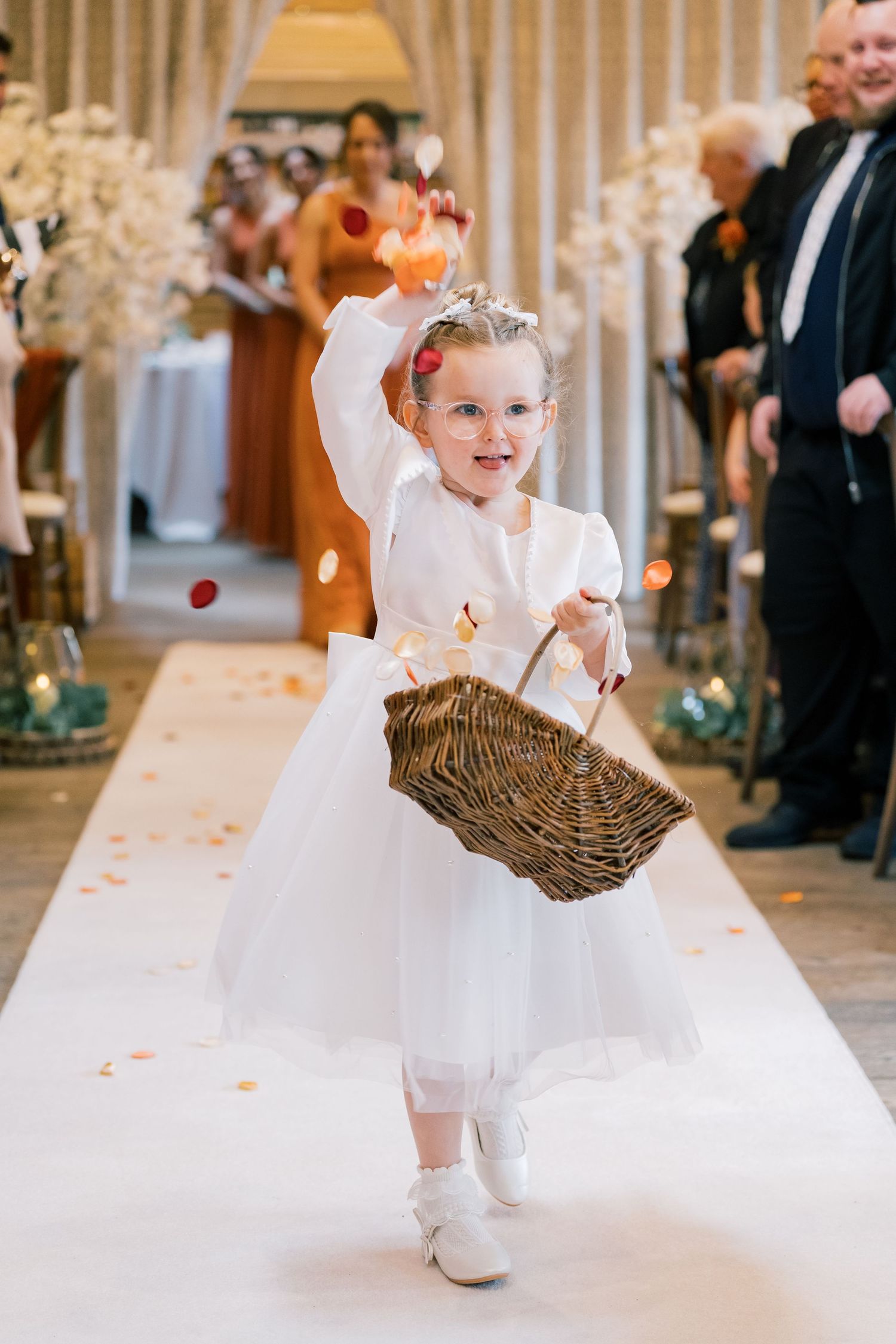 Flower girl in white dress throwing petals while walking down wedding aisle with basket.