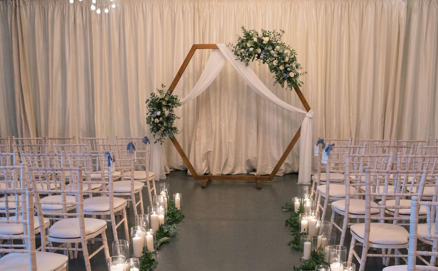 A white draped wedding venue with elegant chandeliers and rows of white chairs set up for a ceremony.