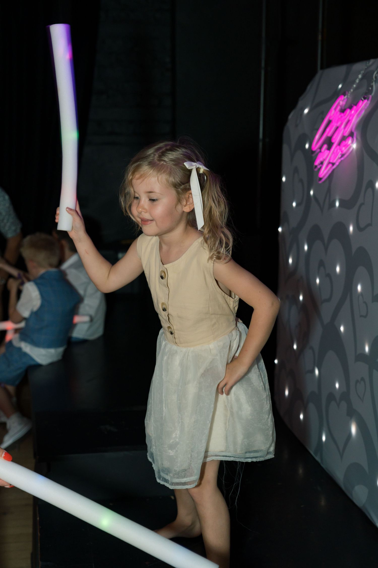 A young flower girl in a beige dress dances under purple party lights at a wedding reception.