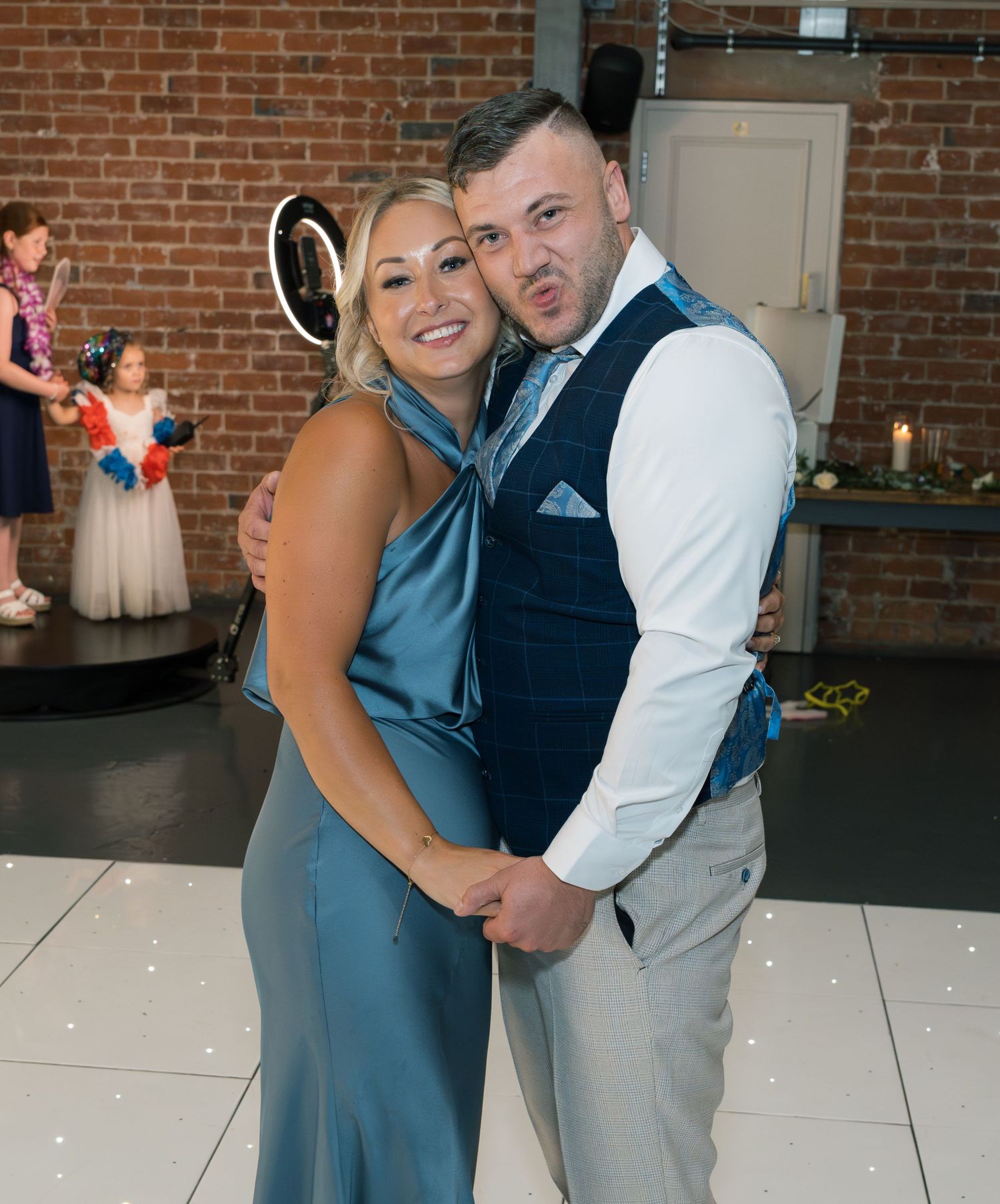 Wedding guests dancing together at reception with brick wall backdrop and white dance floor.