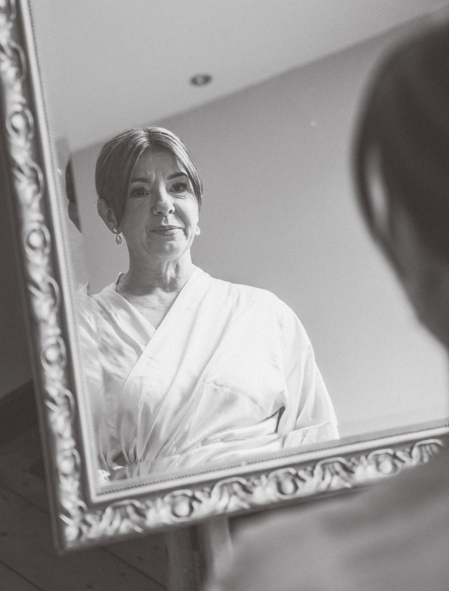 Black and white reflections in an ornate mirror show someone wearing a white robe during wedding preparations.